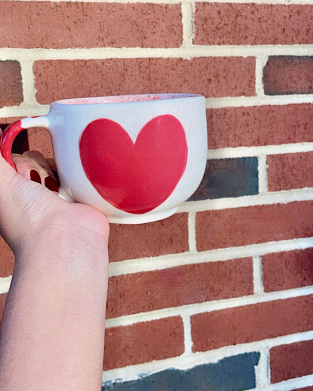 Red heart ceramic mug held in front of a rustic brick wall—an ideal gift for Mother's Day. <3