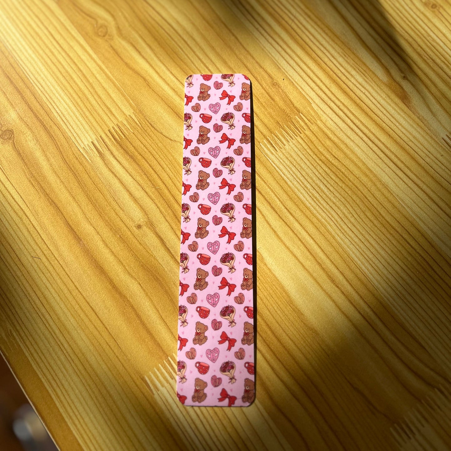A vibrant pink Valentine's Day bookmark featuring cute illustrations of pan concha (Mexican sweet bread), a cute teddy bear, colorful papel picado, and red hearts. The bookmark is designed with love and cheer, making it the perfect addition to any book for a touch of warmth and affection. 💖🍞🧸🎉