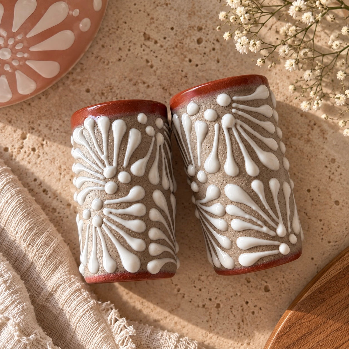 POTTERY SHOT GLASS with a textured beige body, white raised floral patterns, and reddish-brown rims, styled on a speckled beige stone surface with linen and dried flowers.