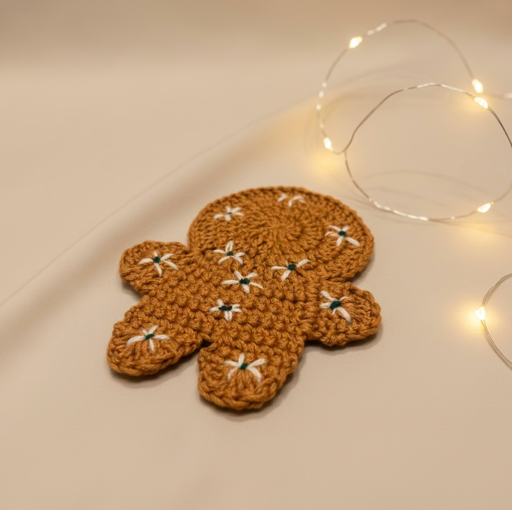 A crocheted gingerbread person-shaped coaster in warm brown, decorated with small white flower motifs and green centers, styled on a beige surface with warm fairy lights in the background.