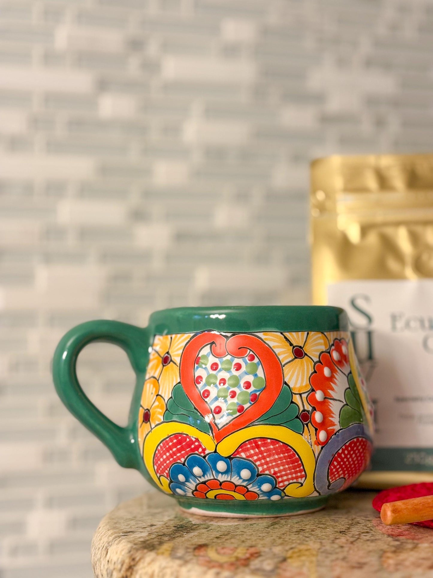 May include: A hand-painted ceramic mug with a green handle and rim. The mug features a colorful floral design with a heart-shaped centerpiece in red, orange, and yellow.