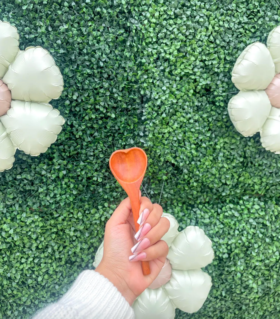 Hand holding a wooden heart-shaped spoon against a green leafy background with decorative flowers.