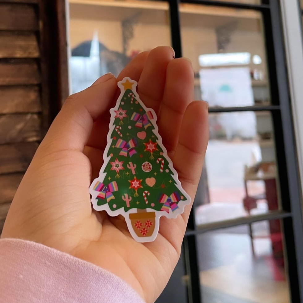 A hand holding a colorful Christmas-themed sticker with a tree and festive patterns.