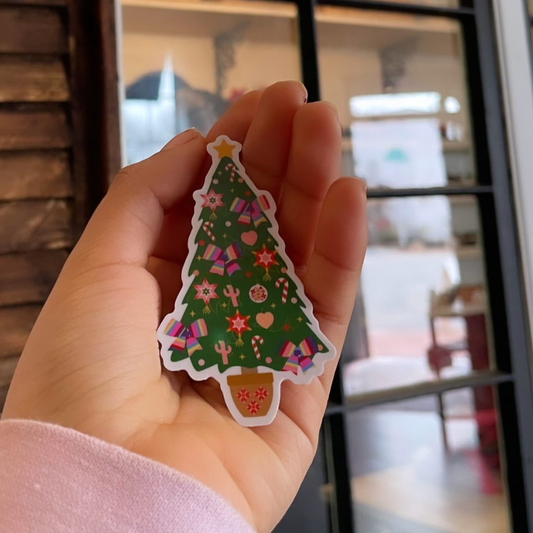 A hand holding a colorful Christmas-themed sticker with a tree and festive patterns.