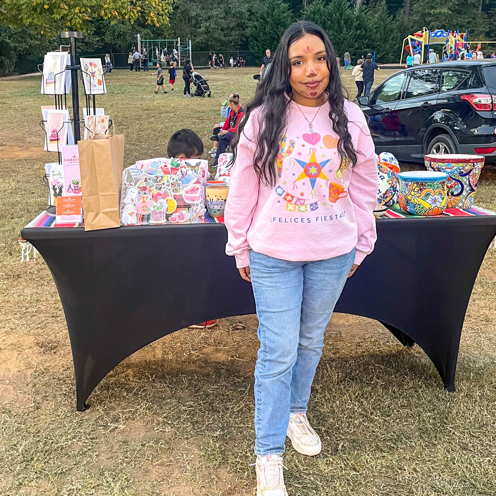 Felices Fiestas sweatshirt in pink at one of our outdoor pop-up events, showcasing the vibrant, hand-drawn design with festive cacti, pan conchas, and piñatas. A perfect combination of style and celebration, captured in a fun, lively outdoor setting.