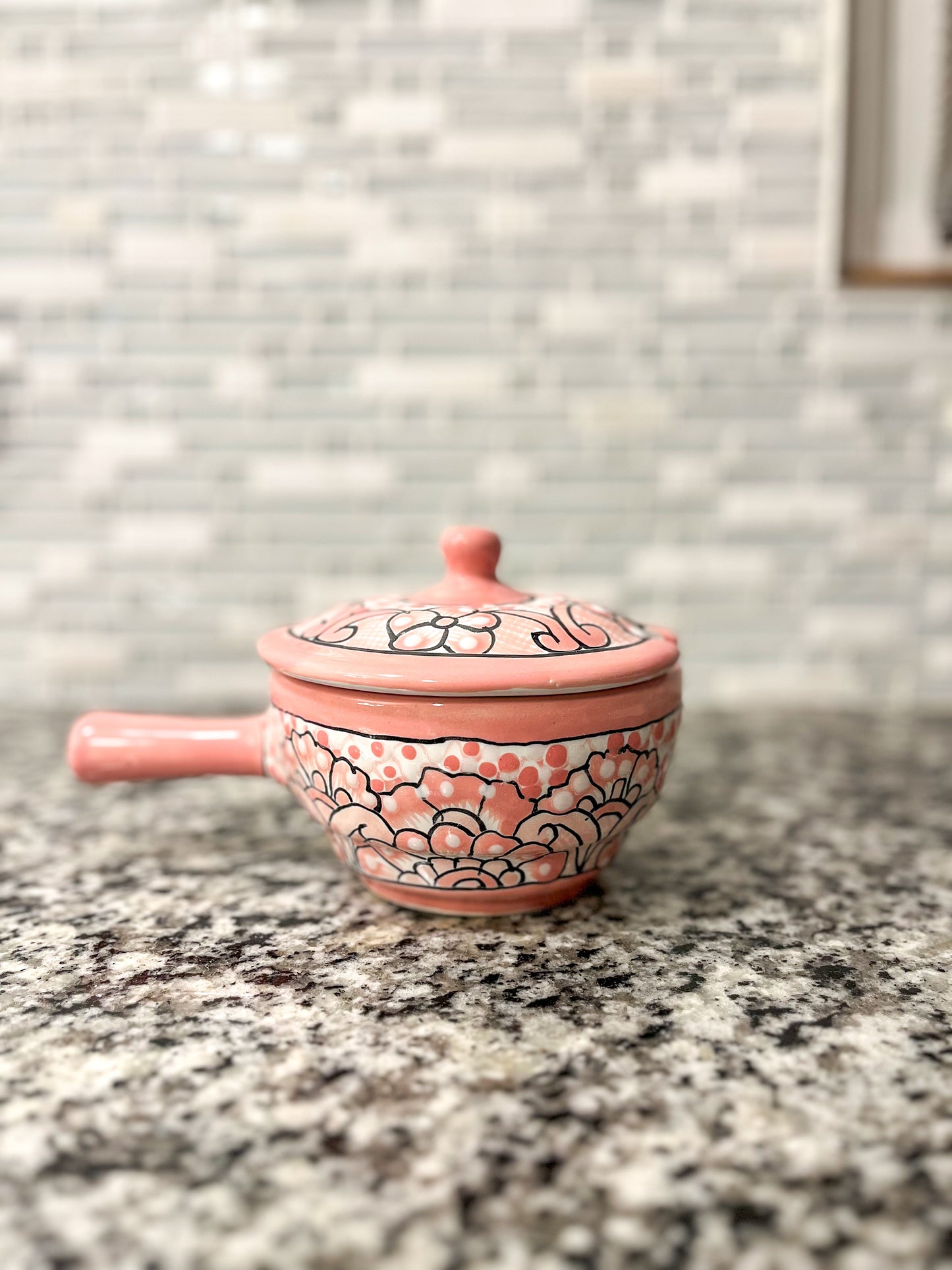 Handmade pink Talavera bowl with lid, elegantly displayed on a granite gray countertop. The bowl features vibrant floral designs and is surrounded by a stylish kitchen setting, including a beautifully patterned tile wall that complements the bowl's colors.