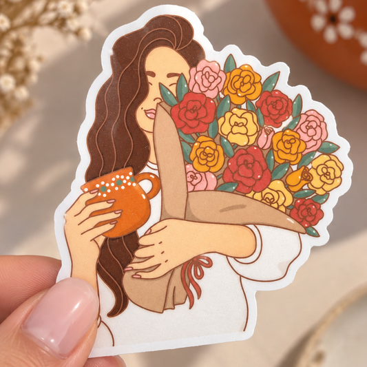 A hand holds a die-cut sticker featuring an illustration of a woman with long brown hair hugging a bouquet of colorful roses while holding an orange mug. The sticker is shown against a softly blurred warm-toned background with dried flowers and a terracotta pot.