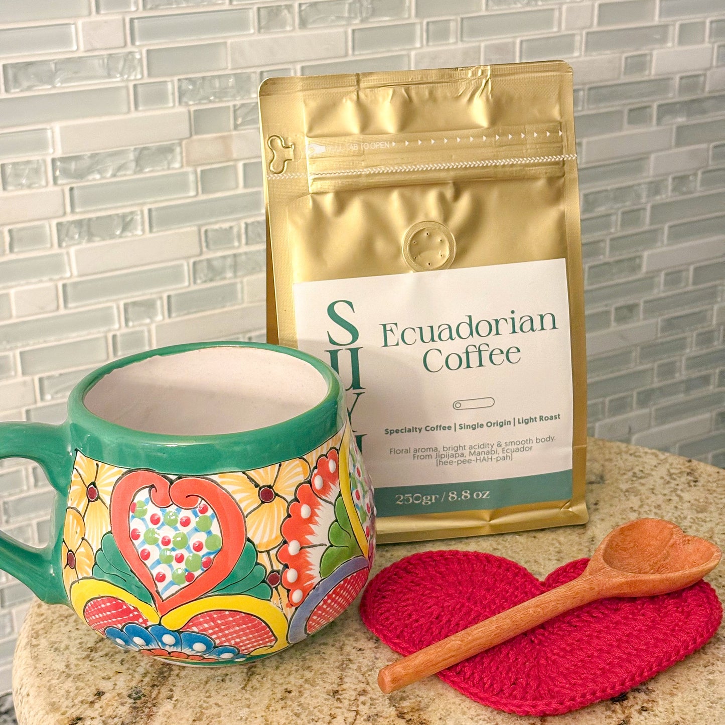 Colorful mug with a heart design, gold coffee package, red coaster, and wooden spoon on a marble surface.