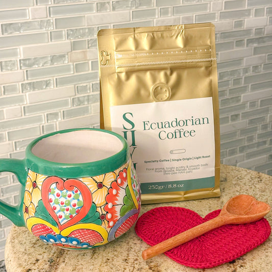Colorful mug with a heart design, gold coffee package, red coaster, and wooden spoon on a marble surface.