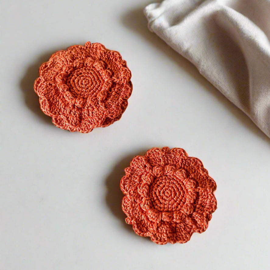 Two orange crochet flowers on a light gray surface with a white fabric bag in the background.