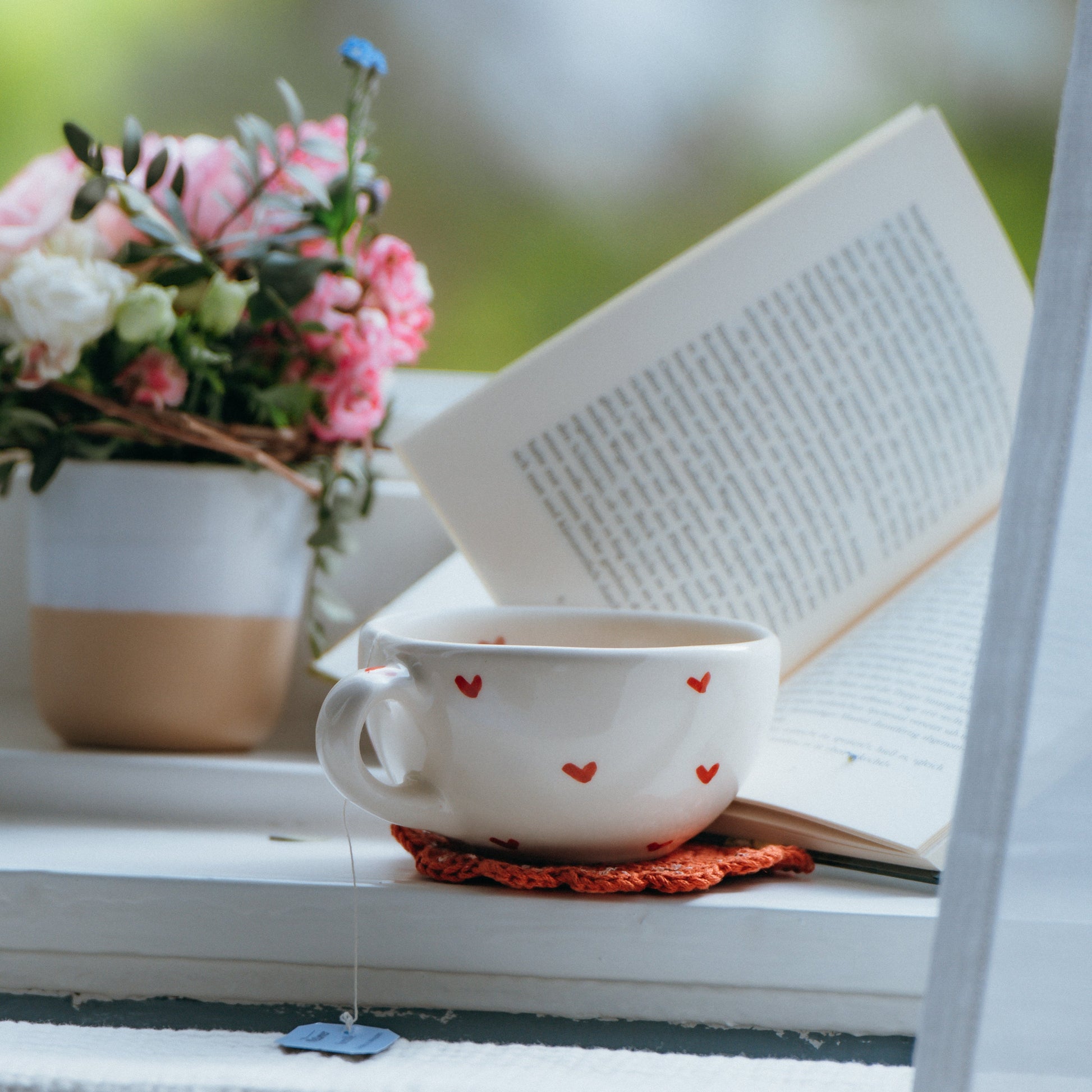 Teacup with heart design on a windowsill next to an open book and small bouquet of flowers.