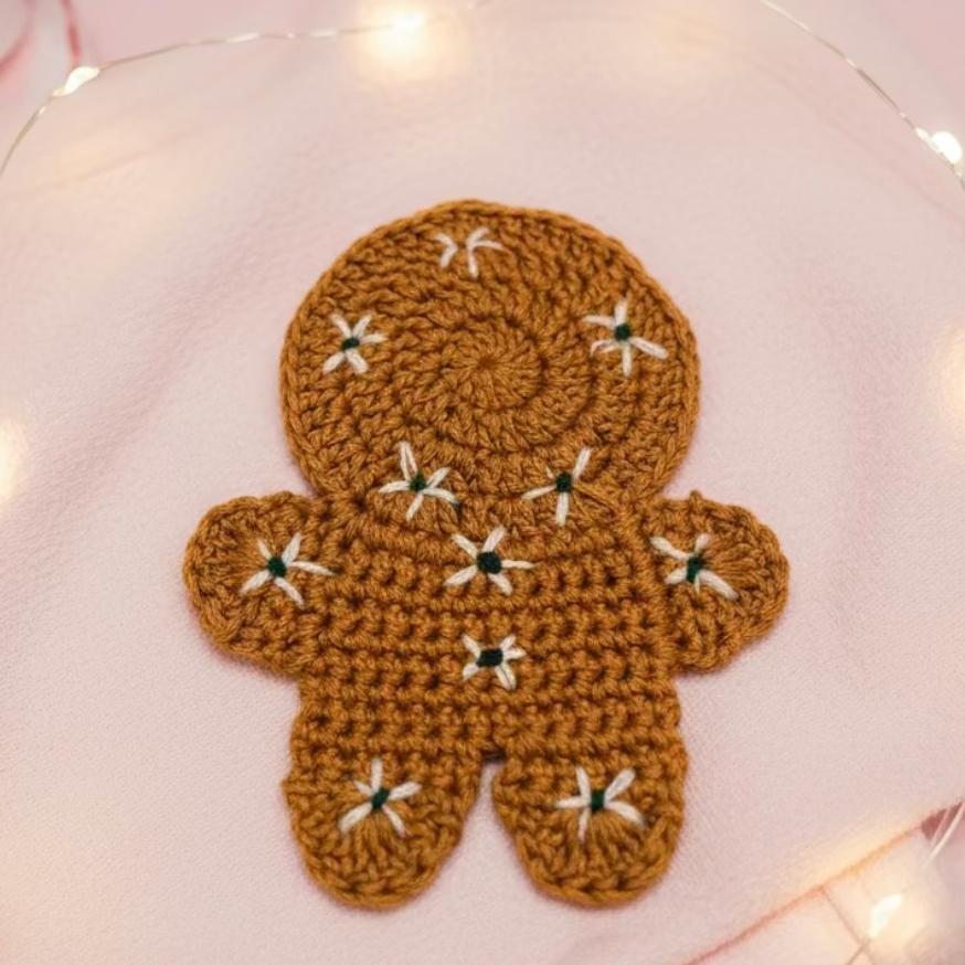 A crocheted gingerbread person ornament in warm brown, decorated with small white embroidered flowers and green centers, resting on a soft pink fabric background with warm fairy lights.
