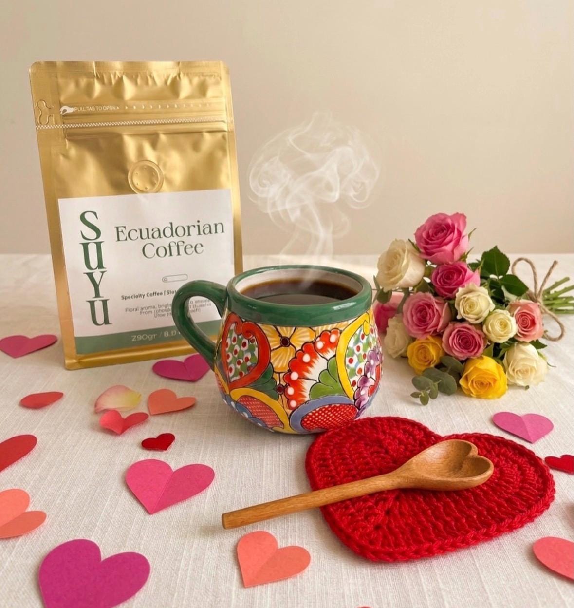 A colorful ceramic mug filled with coffee, a bag of Ecuadorian coffee, and a bouquet of roses. A wooden spoon rests on a red heart-shaped coaster, surrounded by paper hearts. The coffee is steaming.