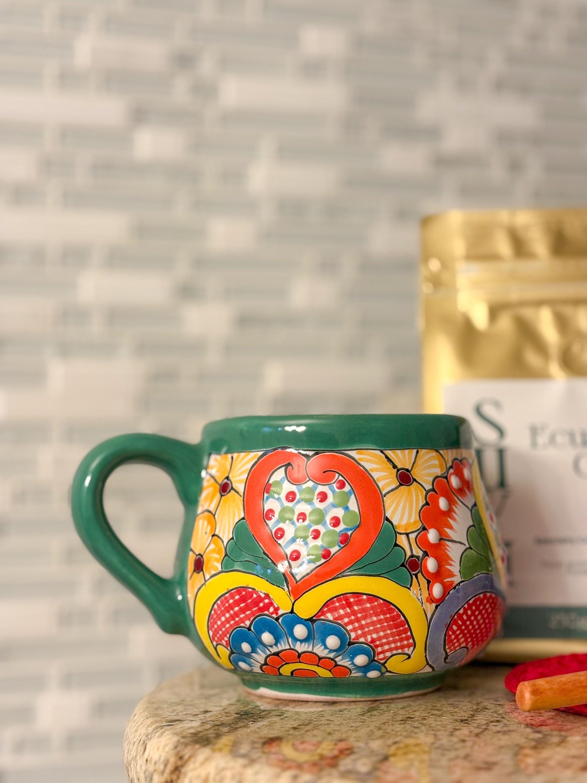 May include: A hand-painted ceramic mug with a green handle and rim. The mug features a colorful floral design with a heart-shaped centerpiece in red, orange, and yellow.