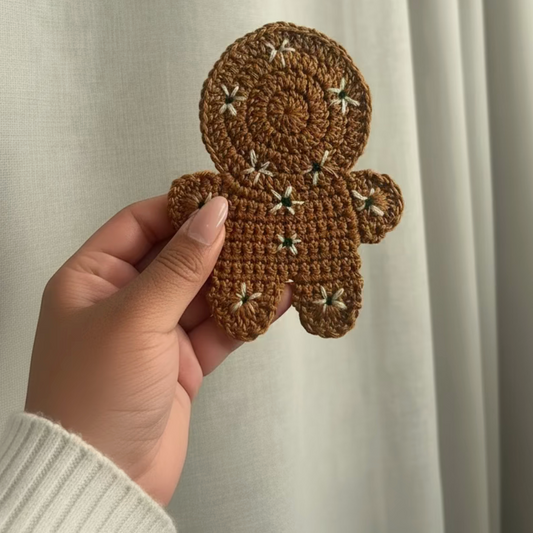 A hand holding a brown crocheted gingerbread person patch with small white embroidered star details, set against a soft beige curtain background.