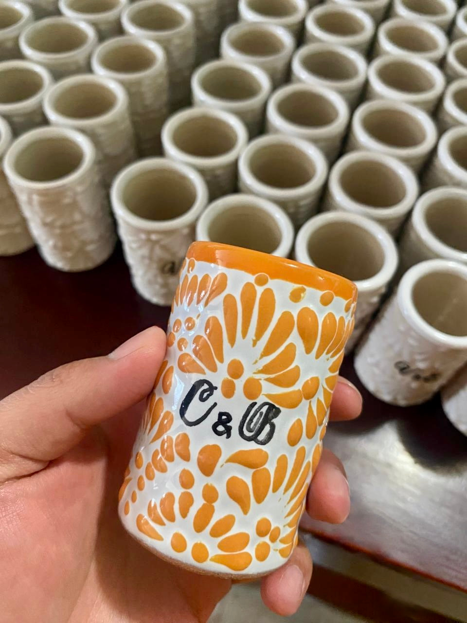Hand holding a ceramic cup with orange pattern and 'C&B' branding, with rows of similar cups in the background.