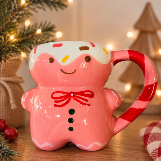 A pink gingerbread person ceramic mug with a white frosted top, red bow, and a candy cane handle, set on a wooden table with warm holiday lights and pine branches in the background.