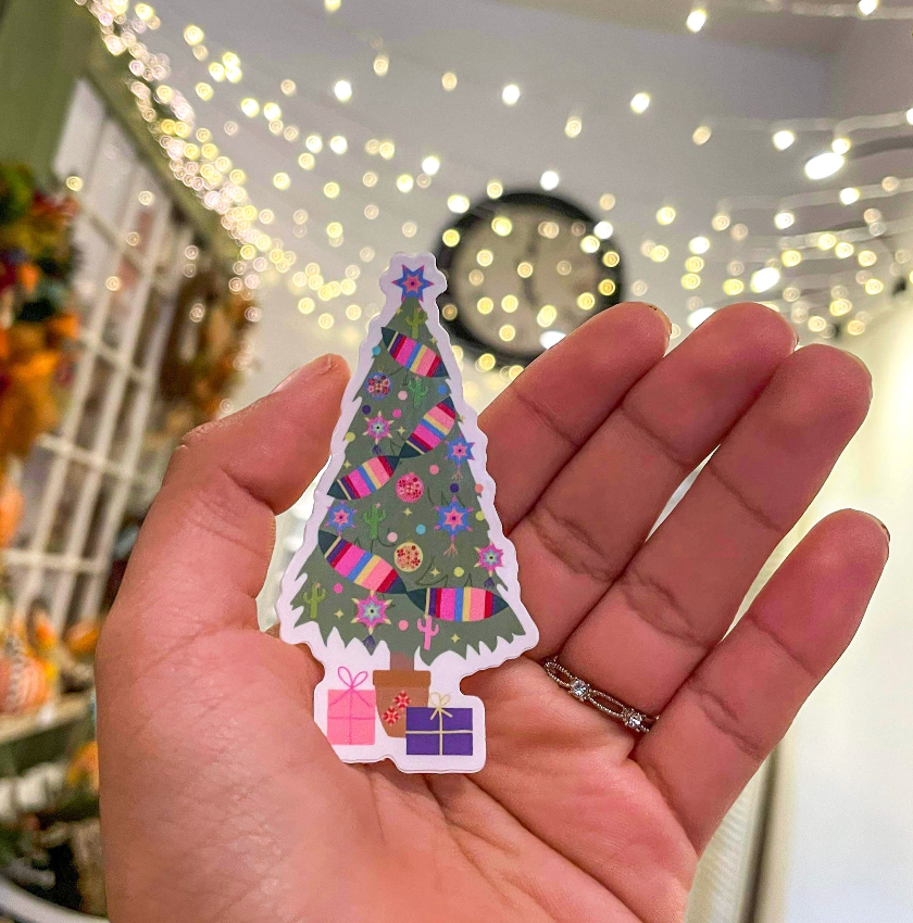 A hand holds a small Christmas tree-shaped sticker or charm decorated with colorful presents, stars, and ornaments, with a blurred indoor holiday background featuring warm string lights.