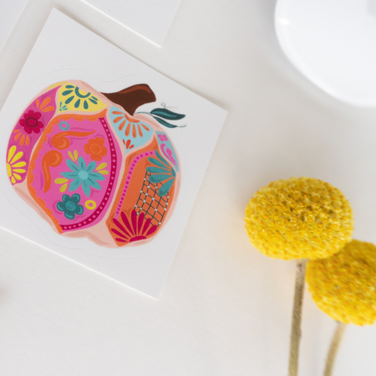 A vibrant pink sticker with a traditional talavera design, featuring a stylized pumpkin, placed on a white surface next to a yellow dried flower.